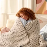 Frau mit lockigem Haar eingekuschelt in einer grob gestrickten sandelholzfarbenen Gewichtsdecke auf einem Sofa, lächelnd und entspannt.