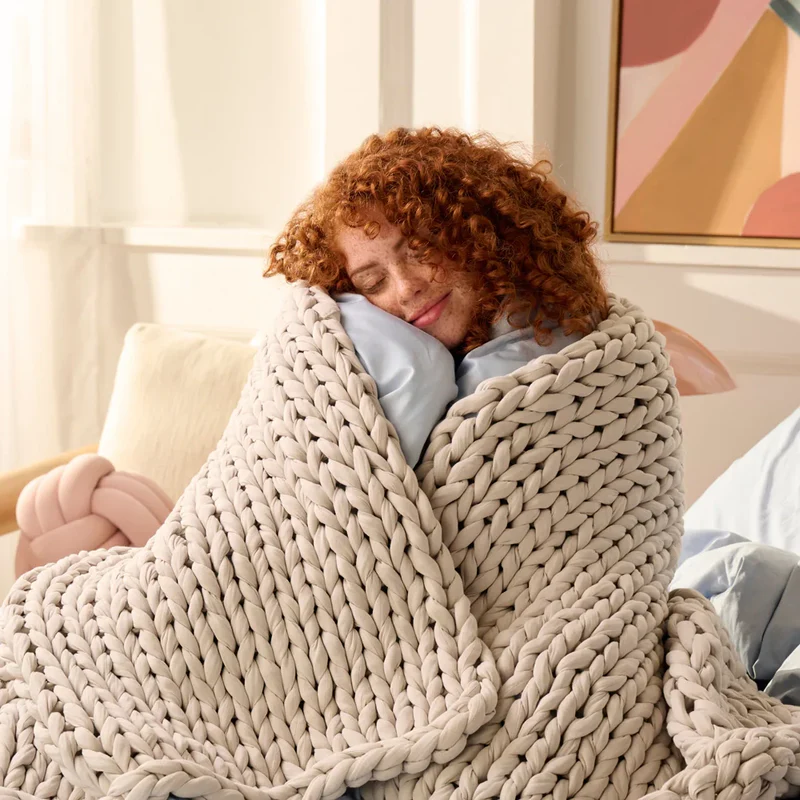 Frau mit lockigem Haar eingekuschelt in einer grob gestrickten sandelholzfarbenen Gewichtsdecke auf einem Sofa, lächelnd und entspannt.