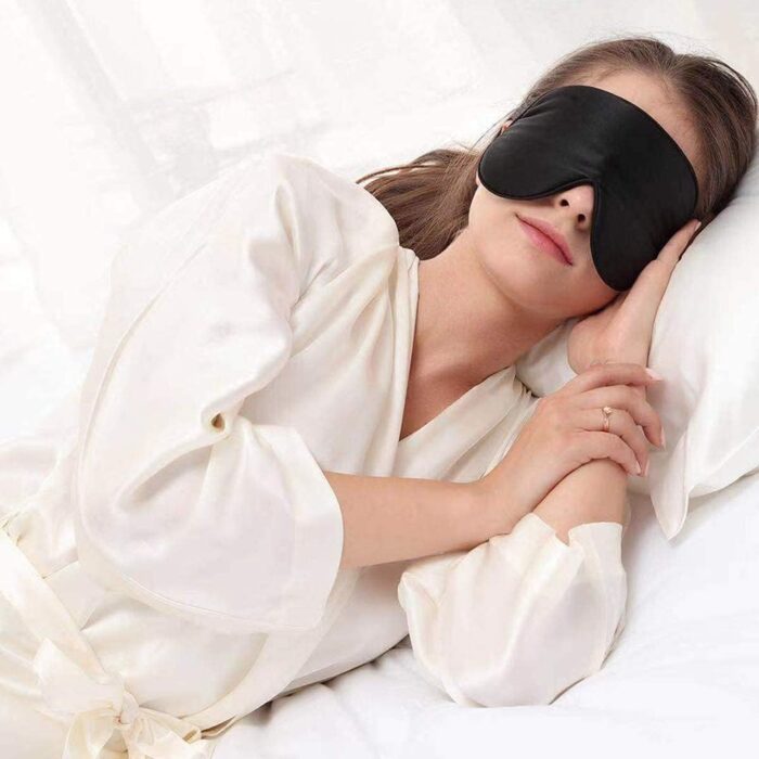 Frau entspannt auf einem Bett und trägt eine schwarze Seiden-Schlafmaske, ideal für erholsamen Schlaf.