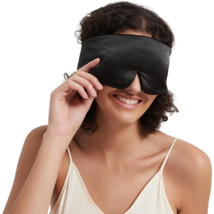 Frau lächelt, während sie eine schwarze Seiden-Schlafmaske anpasst.