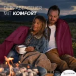 Ein Paar sitzt in der Abenddämmerung am Lagerfeuer, eng aneinander gekuschelt unter einer dunkelroten Decke. Die Frau hält eine Tasse mit der Aufschrift „Mountain Time“, während sie gemeinsam in die Ferne blicken. Im Hintergrund sind Berge und eine wolkige Abendstimmung zu sehen.