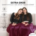 Zwei Frauen sitzen entspannt auf einem Sofa und teilen sich eine grosse, dunkelrote, weiche Reisedecke. Die Decke ist atmungsaktiv, waschmaschinengeeignet und fusselt nicht. Im Hintergrund ein helles Wohnzimmer mit grossen Fenstern.