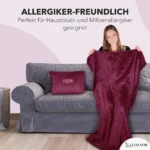 Eine Frau sitzt auf einem grauen Sofa und breitet eine dunkelrote Decke aus. Neben ihr liegt ein kompaktes Kissen mit „Cozyor“-Logo. Text betont die Allergikerfreundlichkeit des Produkts.