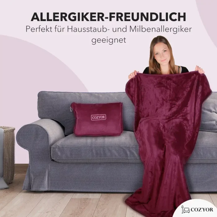 Eine Frau sitzt auf einem grauen Sofa und breitet eine dunkelrote Decke aus. Neben ihr liegt ein kompaktes Kissen mit „Cozyor“-Logo. Text betont die Allergikerfreundlichkeit des Produkts.