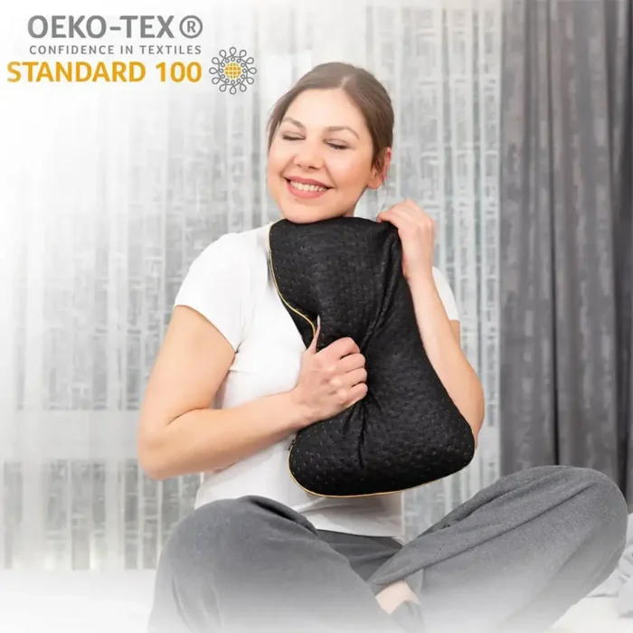 Eine lachende Frau sitzt auf einem Bett und drückt ein schwarzes Memory Foam Reisekissen mit goldener Umrandung an sich. Im Hintergrund sind graue Vorhänge. Oben links ist das OEKO-TEX® STANDARD 100 Siegel zu sehen.