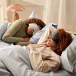 Zwei Personen mit lockigem Haar schlafen entspannt in einem gemütlichen Bett, beide tragen eine gewichtete Schlafmaske in unterschiedlichen Farben. Die Szene vermittelt Ruhe und Erholung in einer entspannten Schlafumgebung.
