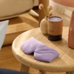 Eine gewichtete Schlafmaske in sanftem Lavendelton liegt auf einem runden Holztisch. Im Hintergrund steht eine dekorative Verpackung mit der Aufschrift "Bearaby The Dreamer", die auf den hochwertigen Inhalt hinweist. Daneben sorgen eine Vase mit Blumen und eine Tischlampe für eine gemütliche Atmosphäre.