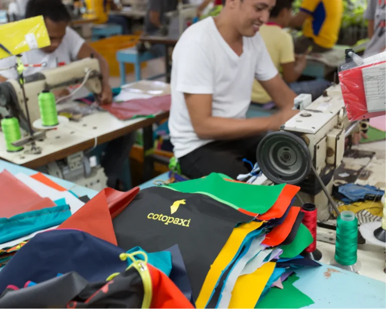 Nähereszene in einer Werkstatt: Mitarbeitende nähen bunte Stoffreste zu Cotopaxi-Taschen zusammen. Im Vordergrund liegen farbenfrohe Stoffzuschnitte mit Cotopaxi-Logo, im Hintergrund sind mehrere Menschen an Nähmaschinen zu sehen.