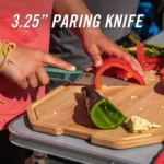 Person schneidet mit einem Camping Messer eine rote Peperoni auf einem Holzbrett, daneben liegen grüne Peperoni und Kerne.