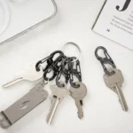 Mehrere Schlüssel sind mit schwarzen Karabiner Schlüsselanhängern an einem Schlüsselring befestigt, daneben liegen ein einzelner Schlüssel mit Karabiner und ein Multitool auf hellem Untergrund.