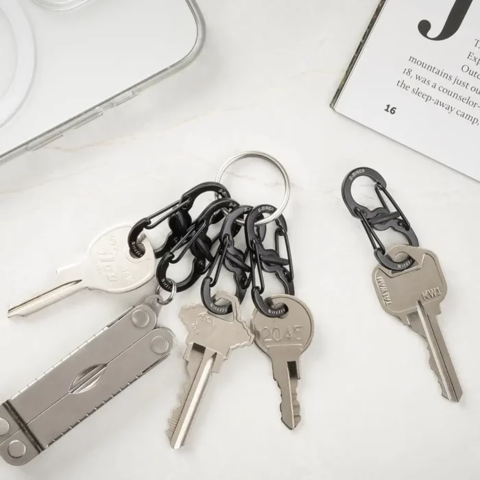 Mehrere Schlüssel sind mit schwarzen Karabiner Schlüsselanhängern an einem Schlüsselring befestigt, daneben liegen ein einzelner Schlüssel mit Karabiner und ein Multitool auf hellem Untergrund.