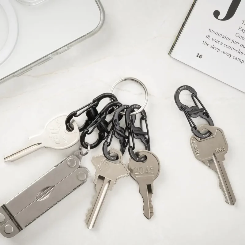 Mehrere Schlüssel sind mit schwarzen Karabiner Schlüsselanhängern an einem Schlüsselring befestigt, daneben liegen ein einzelner Schlüssel mit Karabiner und ein Multitool auf hellem Untergrund.