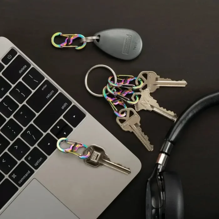 Mehrere Schlüssel und ein Schlüsselanhänger mit Regenbogenfarben-Karabiner Schlüsselanhängern liegen auf einem schwarzen Tisch neben einem Laptop und einem Kopfhörer.