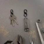 Zwei schwarze Karabiner Schlüsselanhänger auf einer Werkbank – einer hält mehrere Schlüssel, der andere ein Multitool, umgeben von Werkzeugen und Holzspänen.