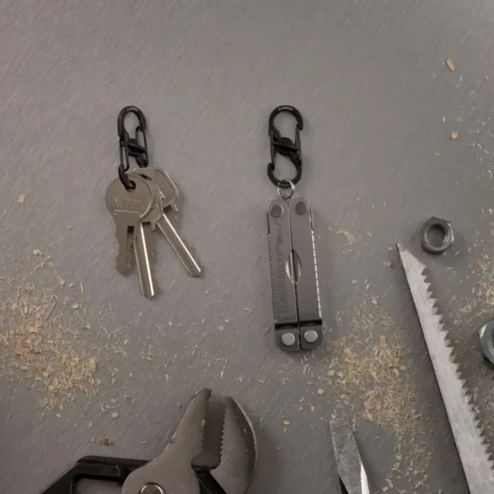 Zwei schwarze Karabiner Schlüsselanhänger auf einer Werkbank – einer hält mehrere Schlüssel, der andere ein Multitool, umgeben von Werkzeugen und Holzspänen.