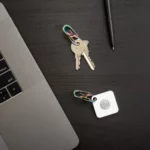 Zwei Karabiner Schlüsselanhänger in Regenbogenoptik liegen auf einem dunklen Schreibtisch – einer mit Schlüsseln, der andere mit einem Tile-Tracker, daneben ein Laptop und ein Stift.