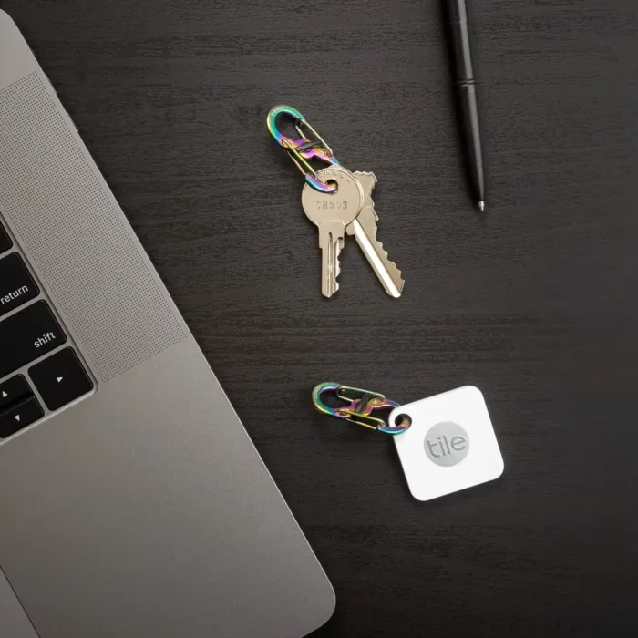 Zwei Karabiner Schlüsselanhänger in Regenbogenoptik liegen auf einem dunklen Schreibtisch – einer mit Schlüsseln, der andere mit einem Tile-Tracker, daneben ein Laptop und ein Stift.