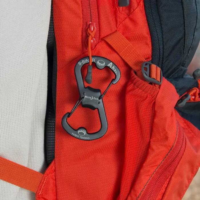 Schwarzer Flaschenöffner Schlüsselanhänger in Karabinerform ist an einer Schlaufe eines roten Rucksacks befestigt.