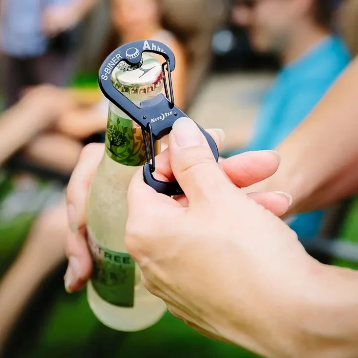 Person öffnet eine Glasflasche mit einem schwarzen Flaschenöffner Schlüsselanhänger in Karabinerform, im Hintergrund sitzen unscharf mehrere Menschen.
