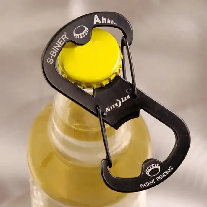Schwarzer Flaschenöffner Schlüsselanhänger in Karabinerform wird an einem gelben Kronkorken einer Glasflasche angesetzt.