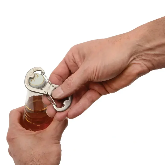 Person öffnet mit einem silbernen Flaschenöffner Schlüsselanhänger in Karabinerform eine Glasflasche mit Kronkorken vor weissem Hintergrund.