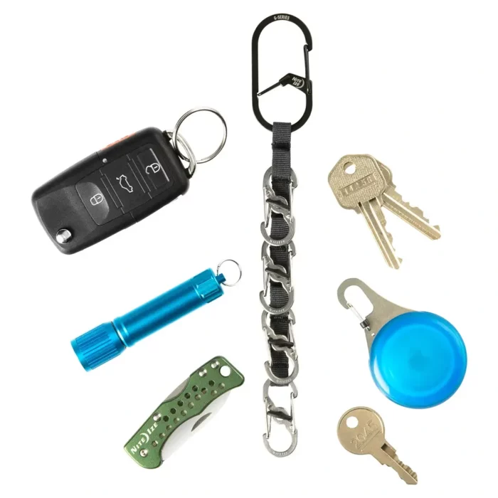 Schlüssel Karabiner mit mehreren kleinen Karabinern an einem schwarzen Band, umgeben von Autoschlüssel, Taschenlampe, Messer, Schlüsseln und AirTag-Halterung vor weissem Hintergrund.