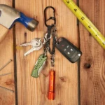 Schlüssel Karabiner mit mehreren befestigten Gegenständen wie Autoschlüssel, Taschenlampe, Taschenmesser und normalen Schlüsseln liegt auf einer Holzfläche neben Hammer, Nägeln und Massband.