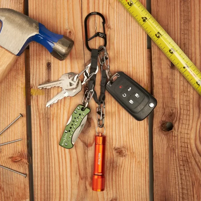 Schlüssel Karabiner mit mehreren befestigten Gegenständen wie Autoschlüssel, Taschenlampe, Taschenmesser und normalen Schlüsseln liegt auf einer Holzfläche neben Hammer, Nägeln und Massband.