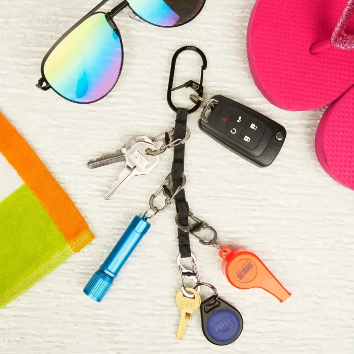 Schlüssel Karabiner mit befestigten Schlüsseln, Autoschlüssel, Taschenlampe, Pfeife und Schlüsselanhänger liegt auf hellem Untergrund neben Sonnenbrille, Flipflops und Badetuch.
