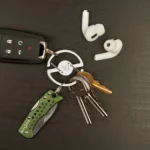 Schlüsselanhänger für Hosenbund mit speziellem Ringmechanismus verbindet mehrere Schlüssel, ein Taschenmesser und einen Autoschlüssel, daneben liegen kabellose In-Ear-Kopfhörer auf dunklem Untergrund.