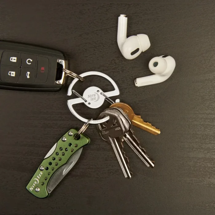 Schlüsselanhänger für Hosenbund mit speziellem Ringmechanismus verbindet mehrere Schlüssel, ein Taschenmesser und einen Autoschlüssel, daneben liegen kabellose In-Ear-Kopfhörer auf dunklem Untergrund.