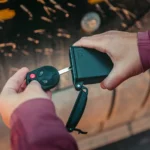 Eine Person legt einen Autoschlüssel in eine schwarze Schlüsselbox für Autoaufbewahrung, im Hintergrund ist ein Fahrzeug unscharf erkennbar.