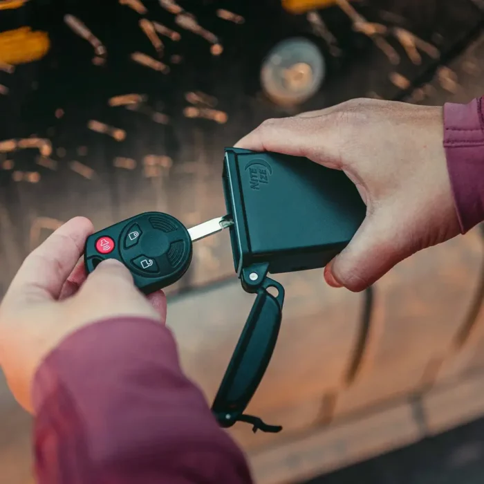 Eine Person legt einen Autoschlüssel in eine schwarze Schlüsselbox für Autoaufbewahrung, im Hintergrund ist ein Fahrzeug unscharf erkennbar.