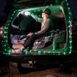 Frau sitzt in einem offenen Kofferraum beim Camping, umgeben von einer grünen Outdoor-LED-Lichterkette und einer Decke, während sie eine Lampe an der Decke befestigt.