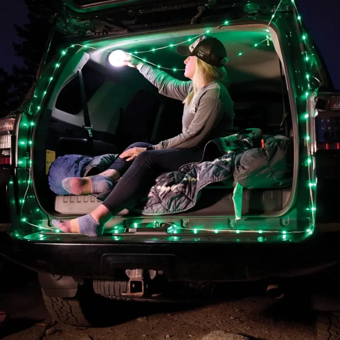 Frau sitzt in einem offenen Kofferraum beim Camping, umgeben von einer grünen Outdoor-LED-Lichterkette und einer Decke, während sie eine Lampe an der Decke befestigt.