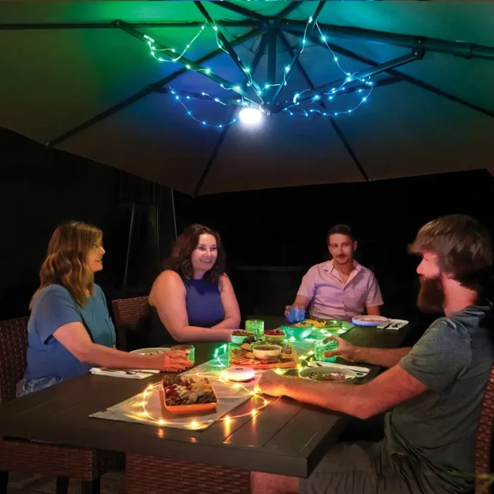 Vier Personen geniessen einen sommerlichen Abend unter einem beleuchteten Sonnenschirm, dekoriert mit farbiger LED-Lichterkette – gemütliche Stimmung beim gemeinsamen Essen im Freien.