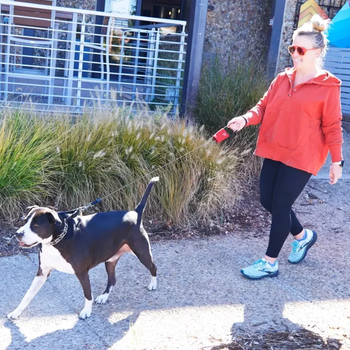Eine Frau spaziert mit einem Hund an einer ausziehbaren Hundeleine – diese ist rot, dünn und fast vollständig ausgezogen.