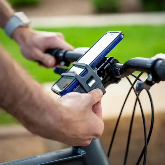 Nahaufnahme einer Handyhalterung am Fahrradlenker. Eine Person befestigt gerade ein Smartphone in der Halterung, die mit flexiblen Gummibändern gesichert ist.