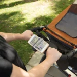 Auf dem Bild ist eine Fahrrad Handyhalterung am Griff eines Kinderwagens befestigt. Ein Smartphone ist darin eingespannt und der Bildschirm ist sichtbar. Die Szene spielt im Freien, auf einem Gehweg neben einer Rasenfläche.