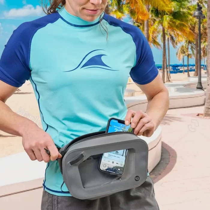 Auf dem Bild steckt eine Person ein Smartphone in eine wasserdichte Gürteltasche mit durchsichtigem Sichtfenster. Die Szene spielt an einem sonnigen Strand mit Palmen und Promenade im Hintergrund.
