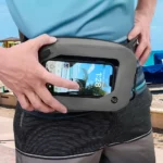 Person bedient ein Smartphone durch das Sichtfenster einer wasserdichten Gürteltasche, im Hintergrund Strandkulisse mit Sonnenschirmen und Meer.