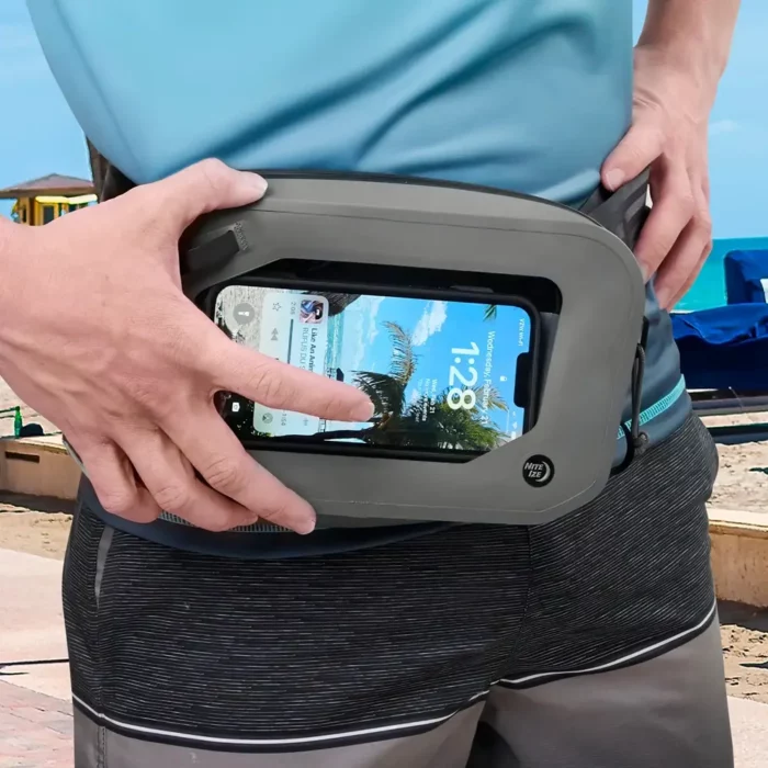 Person bedient ein Smartphone durch das Sichtfenster einer wasserdichten Gürteltasche, im Hintergrund Strandkulisse mit Sonnenschirmen und Meer.