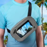 Person trägt eine wasserdichte Gürteltasche quer über die Brust, das Smartphone ist durch das transparente Sichtfenster sichtbar – im Hintergrund tropische Strandkulisse mit Palmen.