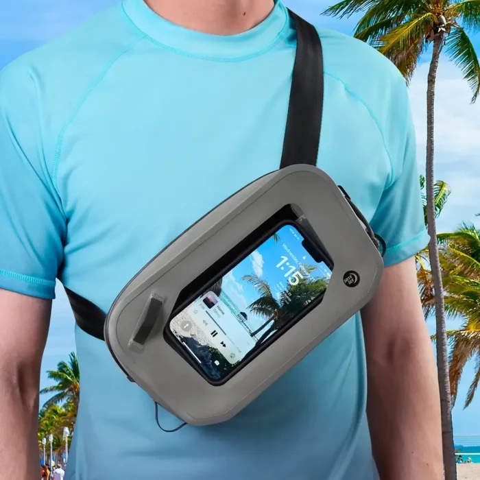 Person trägt eine wasserdichte Gürteltasche quer über die Brust, das Smartphone ist durch das transparente Sichtfenster sichtbar – im Hintergrund tropische Strandkulisse mit Palmen.