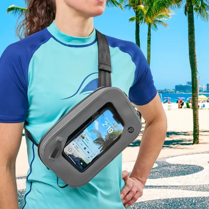 Person trägt eine wasserdichte Gürteltasche quer über die Schulter am Strand – das Smartphone ist durch das transparente Sichtfenster gut bedienbar.