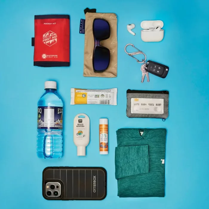 Flach auf einem blauen Untergrund ausgebreitet liegen typische Gegenstände für einen Ausflug – darunter Wasserflasche, Sonnencreme, Sonnenbrille, Erste-Hilfe-Set, Schlüssel und ein Smartphone.