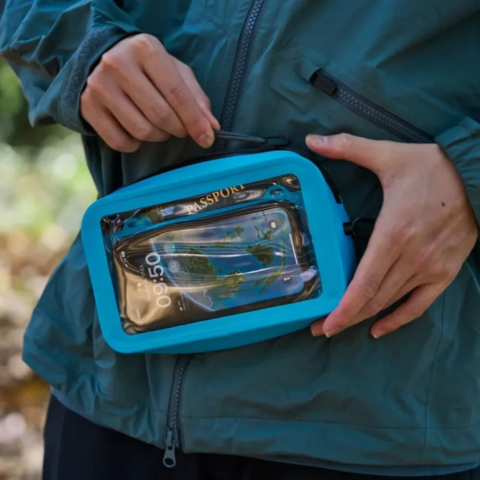 Person mit türkisfarbener Jacke hält eine wasserdichte Bauchtasche in kräftigem Blau, darin gut sichtbar ein Smartphone und ein Reisepass.