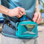 Person verstaut ein Smartphone in einer türkisfarbenen, wasserdichten Bauchtasche mit transparenter Fronttasche.