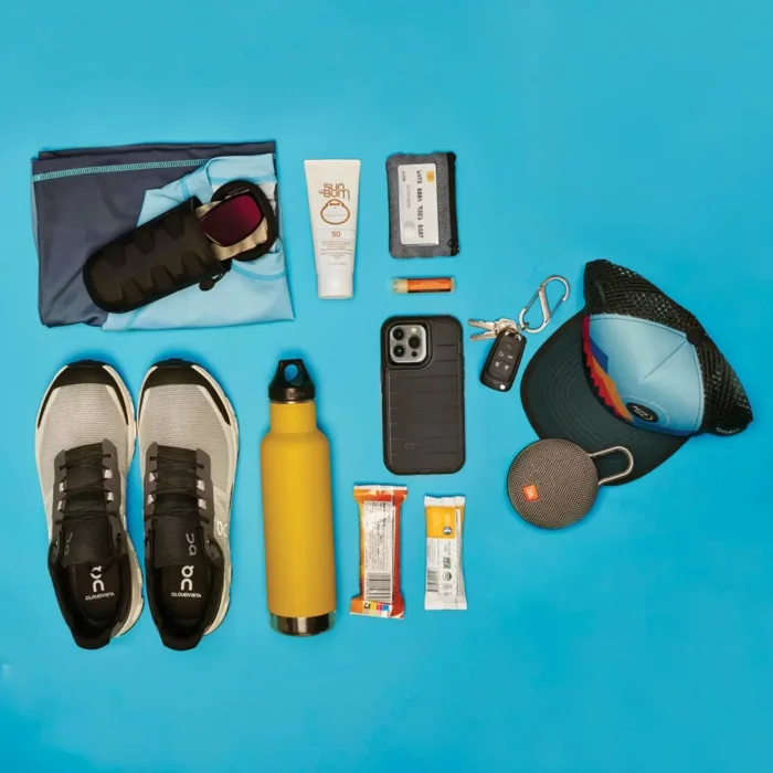 Flatlay auf türkisfarbenem Hintergrund zeigt Essentials für einen aktiven Tag draussen: Laufschuhe, Sportkleidung, Trinkflasche, Sonnencreme, Snacks, Schlüssel, Sonnenbrille, Cap und kompakter Lautsprecher.
