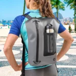 Frau spaziert entspannt am Strandboulevard entlang – auf dem Rücken ein moderner, grauer wasserdichter Rucksack mit Netztasche, aus der eine Edelstahl-Trinkflasche hervorschaut.